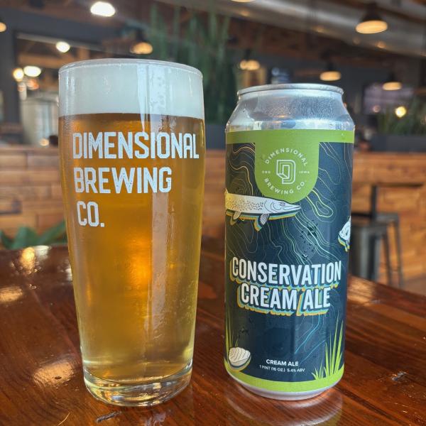 a glass of pale ale with the Dimensional Brewing logo next to a can of conservation cream ale