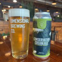 a glass of pale ale with the Dimensional Brewing logo next to a can of conservation cream ale